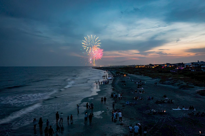 Fireworks light up the twilight sky over a crowded beach, with people gathered along the shoreline watching the display.
