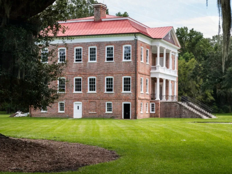 A historic brick mansion with white trim, red roof, multiple windows, and a grassy lawn set among trees and mossy branches.