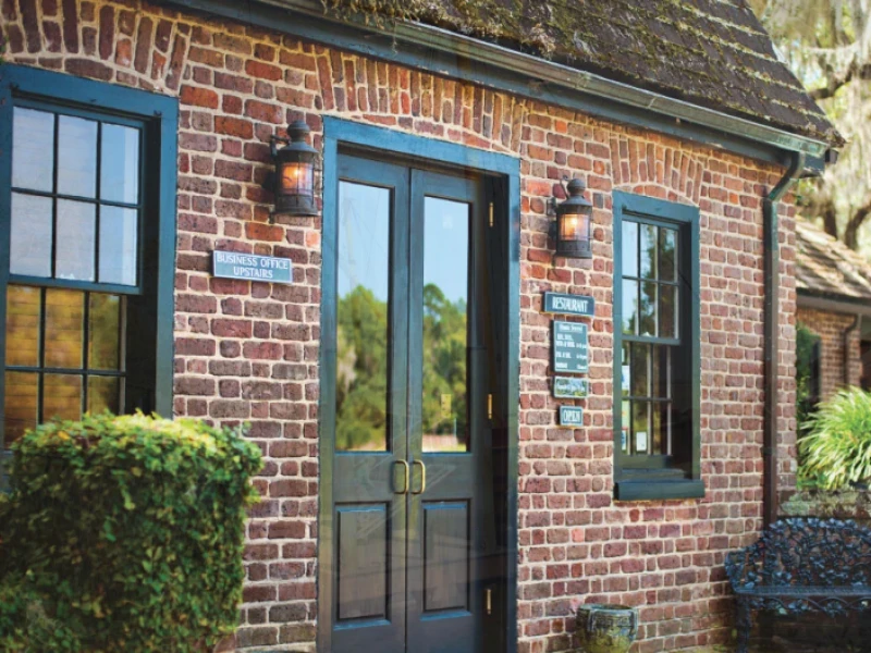 A brick cottage with blue doors and windows, black lanterns, and lush greenery flanking a small entryway.