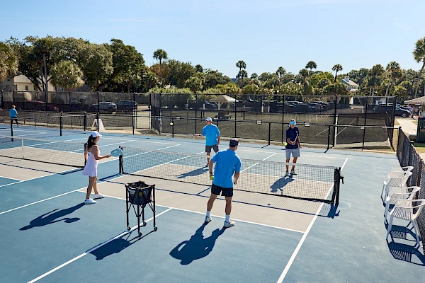 Two people on a tennis court: a man in a green shirt foreground left, a woman in white and teal outfit smiling in background.