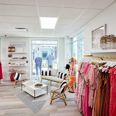 A bright boutique with clothing racks, shelves, and a seating area; colorful dresses on the right, pastel decor, and a front window showing passersby.