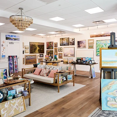 A bright art gallery filled with paintings, prints, and framed works on walls; sofas and tables display decor, with an easel and lamps around. Top it at 140 characters, always ending the sentence.