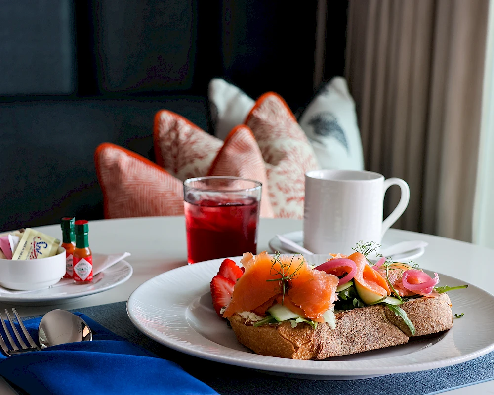 A breakfast table with toast topped with smoked salmon and greens, a glass of red juice, a white cup, and colorful napkins in a cozy, candlelit setting.