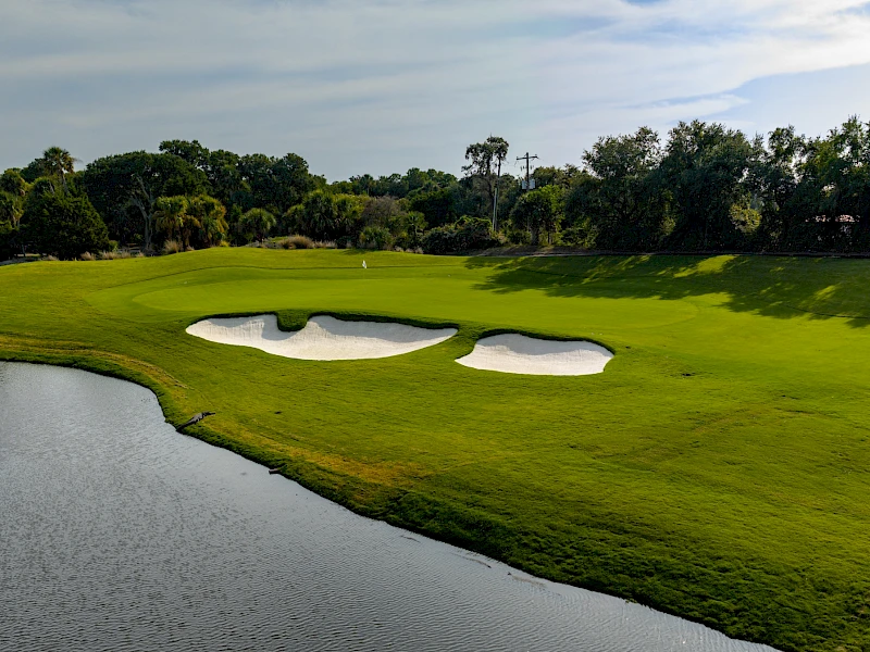 A lush golf course with green fairways, sand bunkers, and a water hazard surrounding the greens, under a partly cloudy sky.
