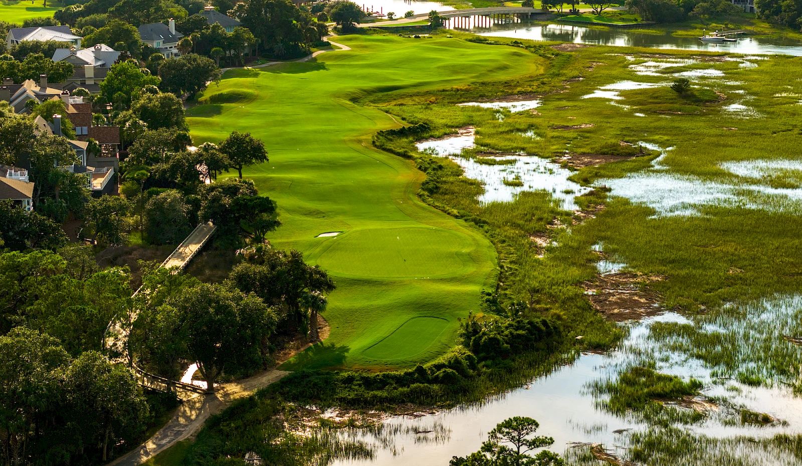 An aerial view of a lush golf course winding through green fairways, bordered by trees and water, with residential areas along the left edge.
