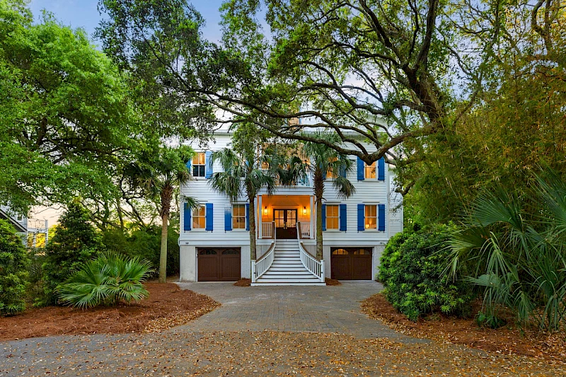 A charming blue two-story house with white trim, surrounded by big trees and a curved front walkway leading to a double garage.