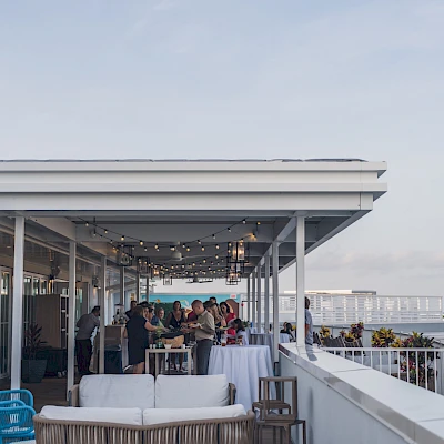People dining on a rooftop terrace with string lights, white couches, and ocean views in the background, enjoying a social gathering.