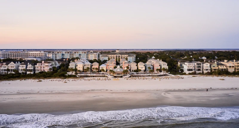 A tranquil seaside scene with a sandy beach in the foreground, gentle waves, and a row of pastel houses along the shore at sunset.