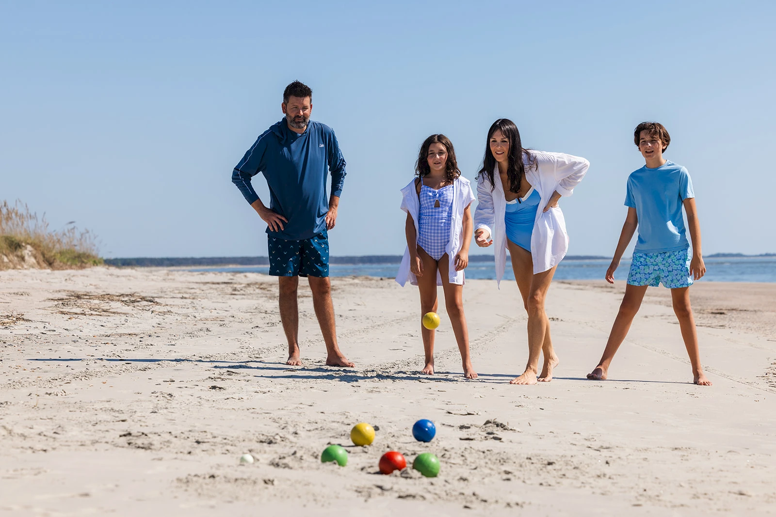 Four people—two adults and two kids—on a sunny beach play bocce or lawn bowls with colorful balls scattered on the sand.