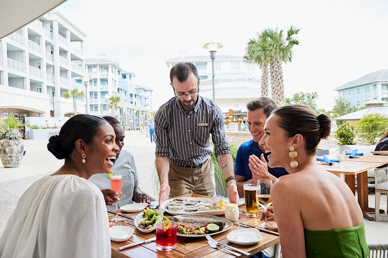 A group of friends enjoys a sunny outdoor meal by the pool, sharing food and drinks while chatting happily in smart casual attire.