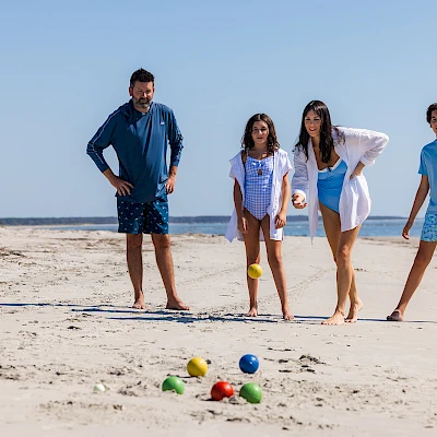 A family of four on a sunny beach play bocce/petanque with colorful balls scattered on dry sand, sea in background, everyone watching.