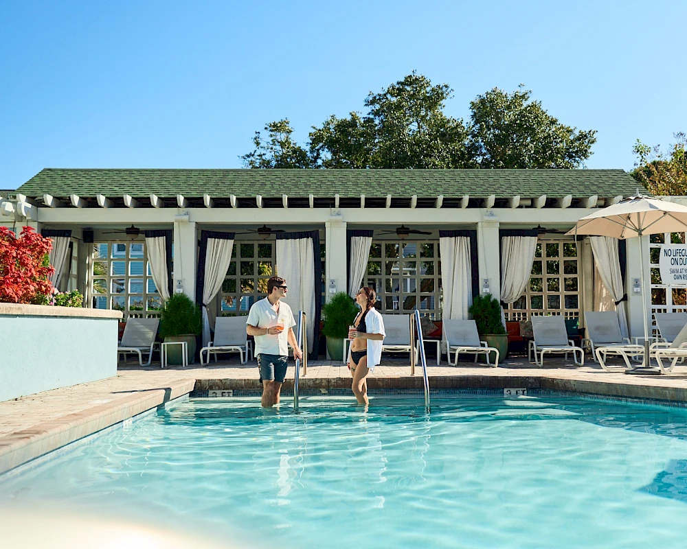 Two people stroll into a sunny pool area of a resort, leaning casually by the water as lounge chairs and umbrellas line the deck.