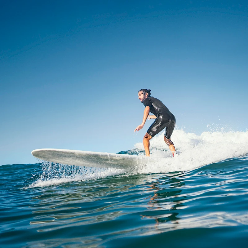 A person stands on a surfboard riding a small wave, gliding across blue ocean under a clear sky.