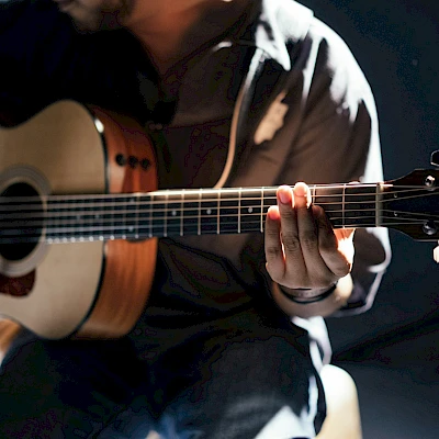 A person strums an acoustic guitar, close-up on the neck and hand, in a dimly lit setting.