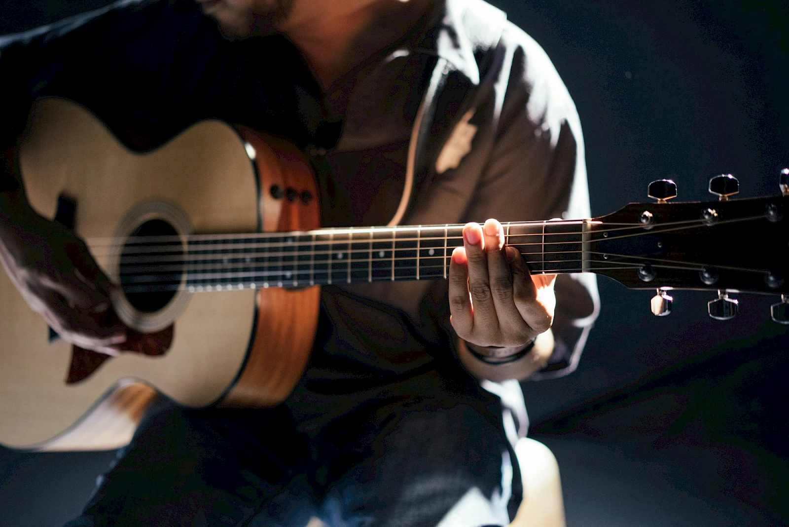 A person sits playing an acoustic guitar, focusing on strumming the strings in a dim, moody setting.