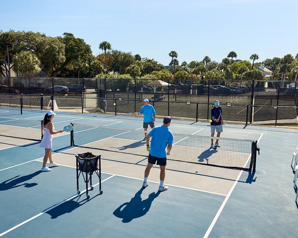 People playing a game of pickleball on outdoor courts, with a net, paddles, and spectators nearby, under a sunny sky.