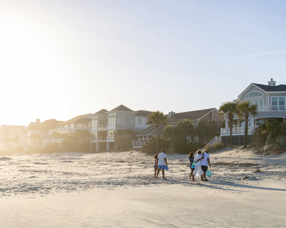 A sunny beach scene with a few people walking along the shore near seaside houses, gentle waves and bright sky, relaxing coastal vibes.