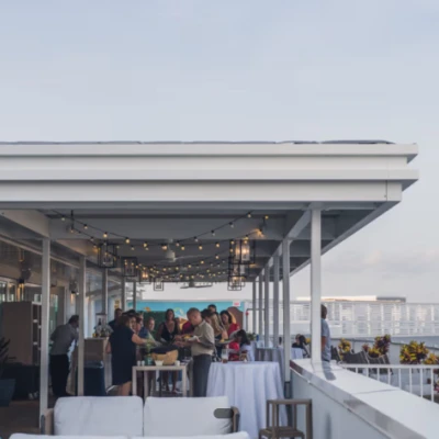 A rooftop party scene with people mingling under string lights, tables set for a gathering, and a view of buildings and the ocean in the background.