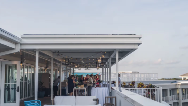 A rooftop party scene with people mingling under string lights, tables set for a gathering, and a view of buildings and the ocean in the background.