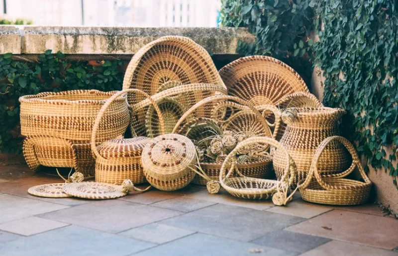 A pile of woven wicker baskets of various sizes stacked outdoors on a tiled patio, some round and curved, with greenery in the background.