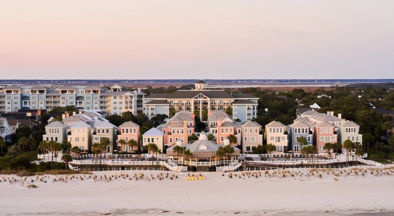 A beachfront resort with multiple pastel-colored villas, white sand, and a calm sea in the foreground, under a pastel sky at dusk.
