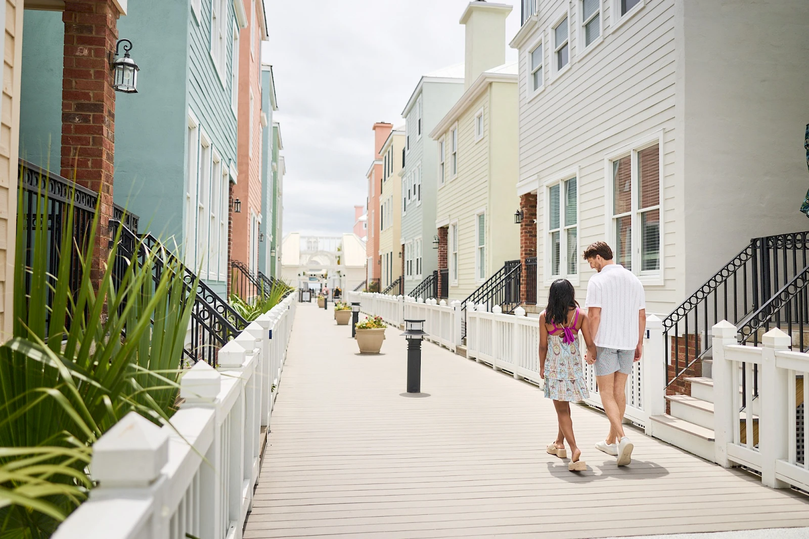 A couple walks along a sunny boardwalk lined with pastel beach houses; bright doors, railings, and potted plants frame the path.