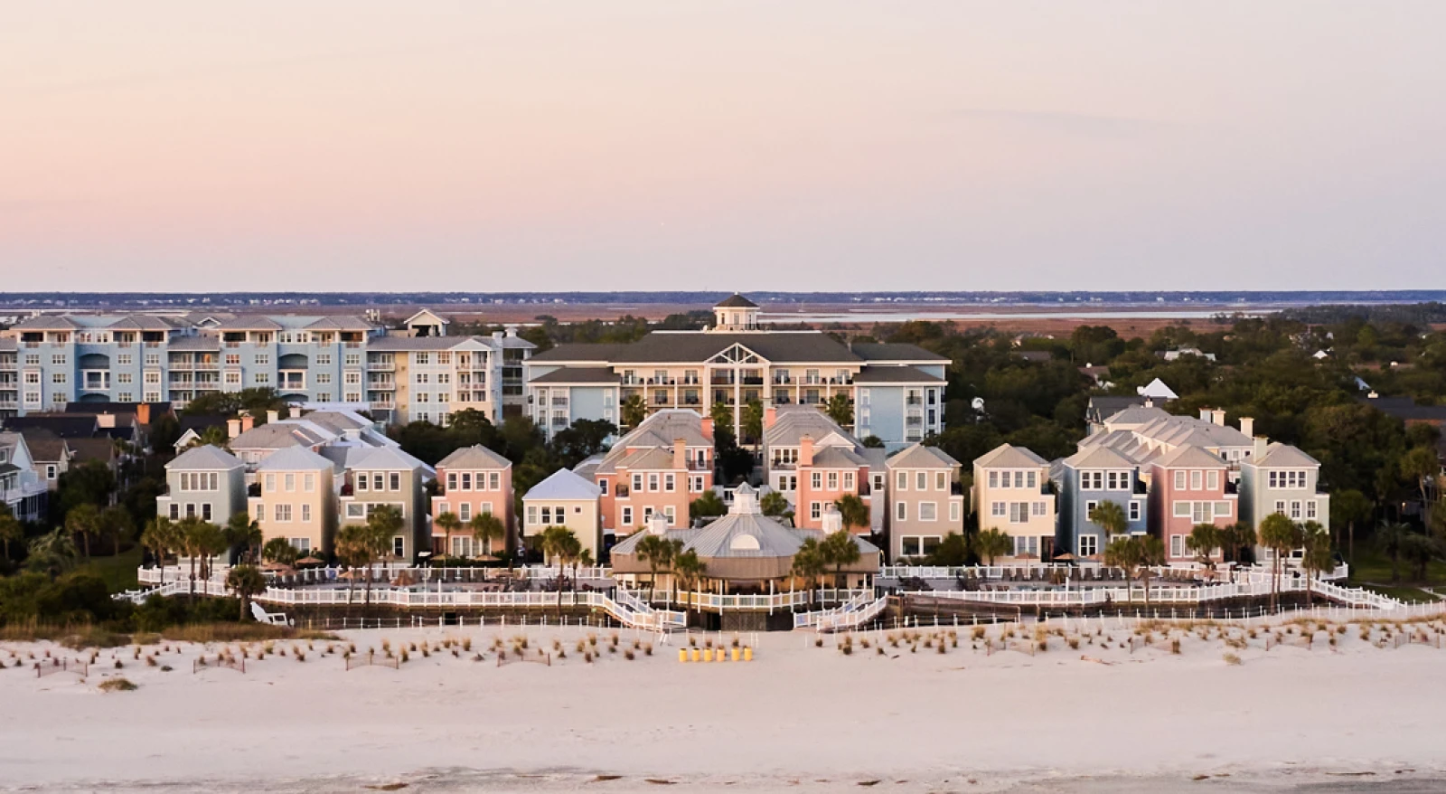 A row of pastel beach houses along a sandy shore, with a calm ocean in front and a low-rise resort feel under a soft sunset sky.