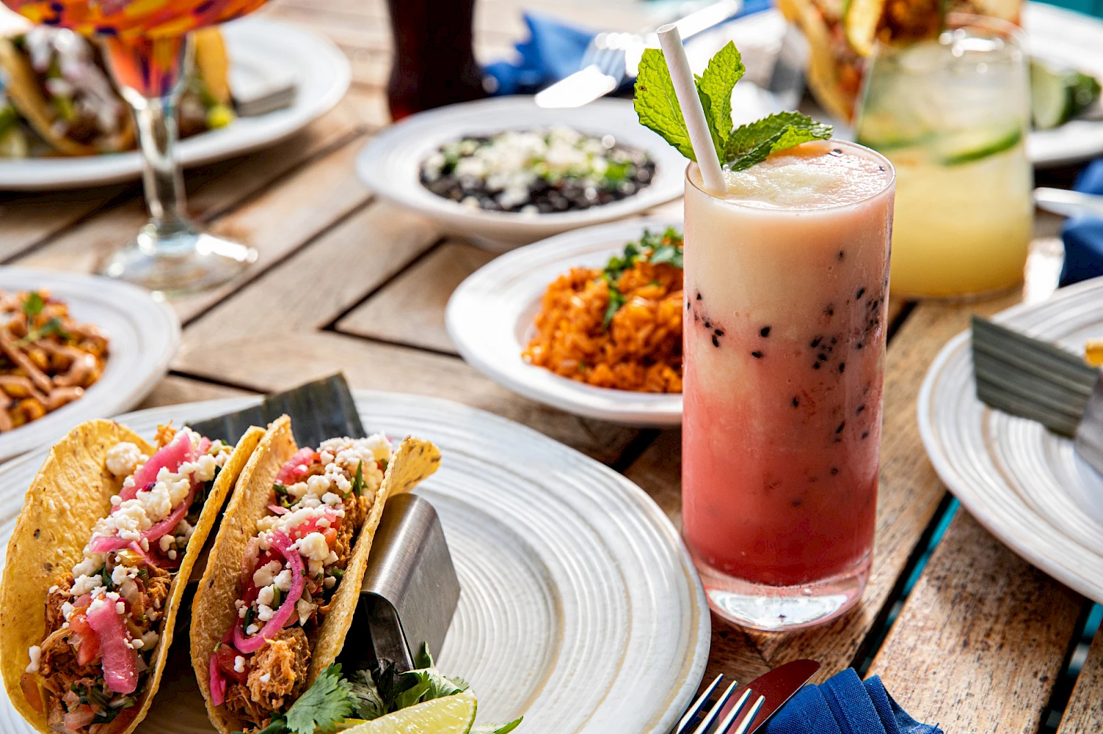 A sunny outdoor meal: tacos with toppers, bowls of sides, a pink fruit drink in a tall glass, and other plates on a wooden table.