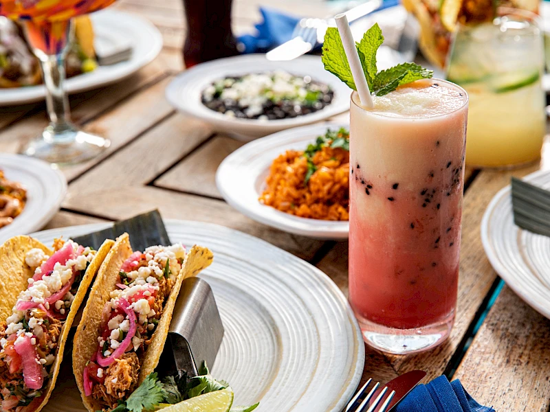 A sunny outdoor meal: tacos with toppers, bowls of sides, a pink fruit drink in a tall glass, and other plates on a wooden table.