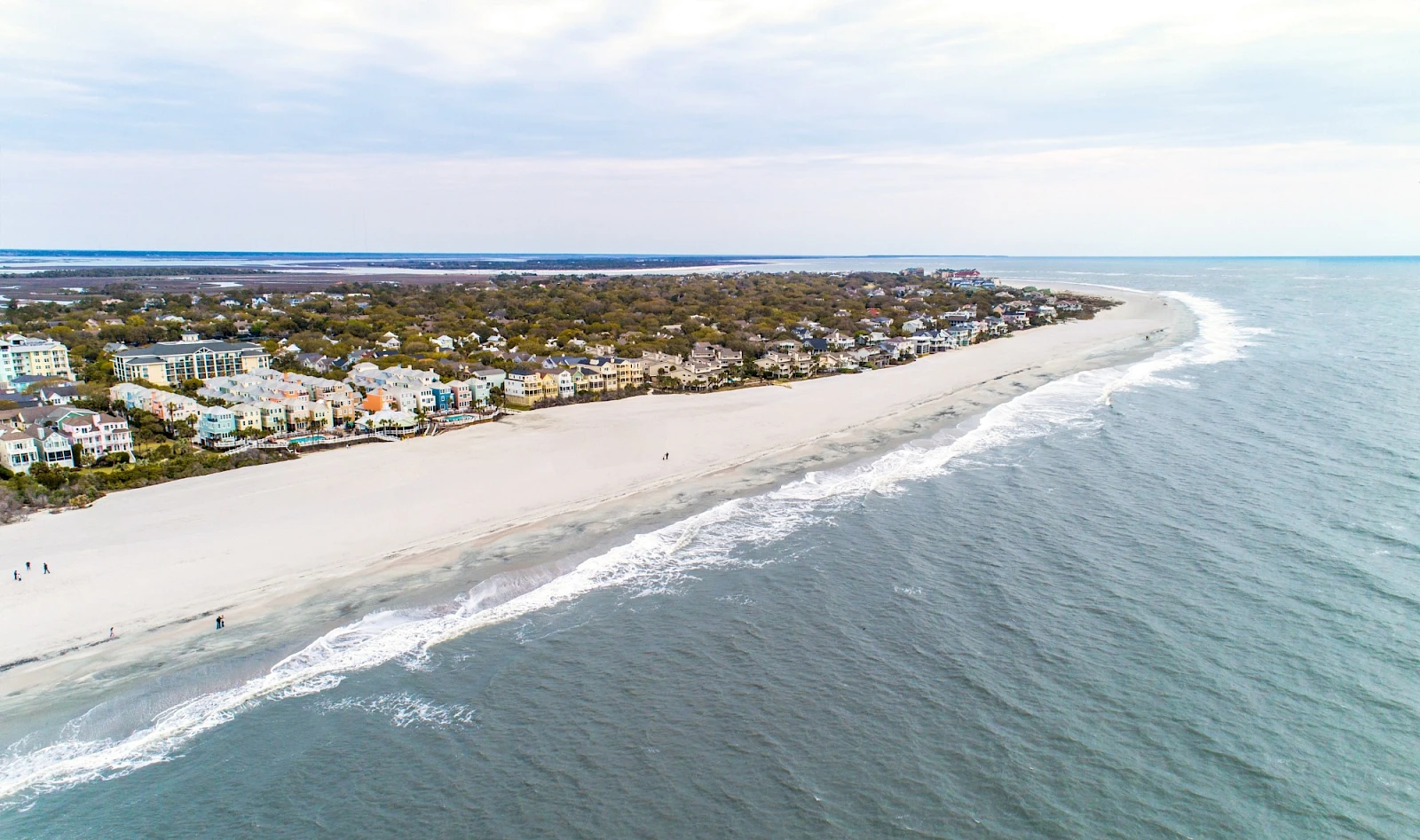 Aerial view of a sandy beach with calm waves, lined by houses and greenery along the coast, stretching into the distance.