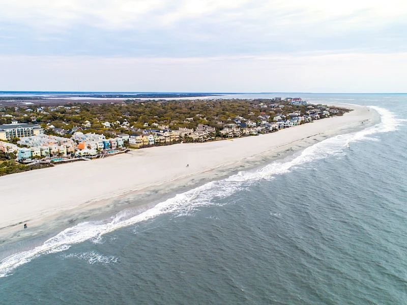 Aerial view of a sandy beach with calm waves, lined by houses and greenery along the coast, stretching into the distance.