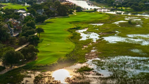 A lush green golf course tucked between water and houses, with bright fairways, trees lining the holes, and a scenic wetland along the edges.