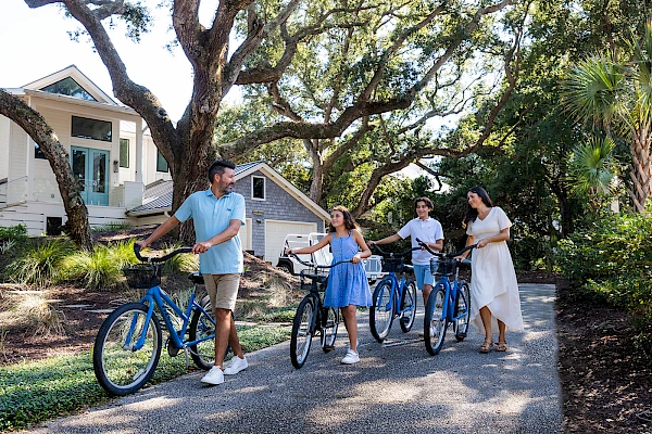 A family bikes along a shaded path near a white house, with two kids and two adults enjoying a sunny day on blue bicycles.