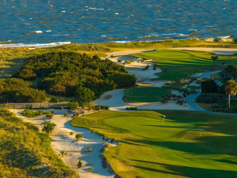 A coastal golf course with green fairways winding along sandy paths, low shrubs, and the sea in the background, sunny and scenic.