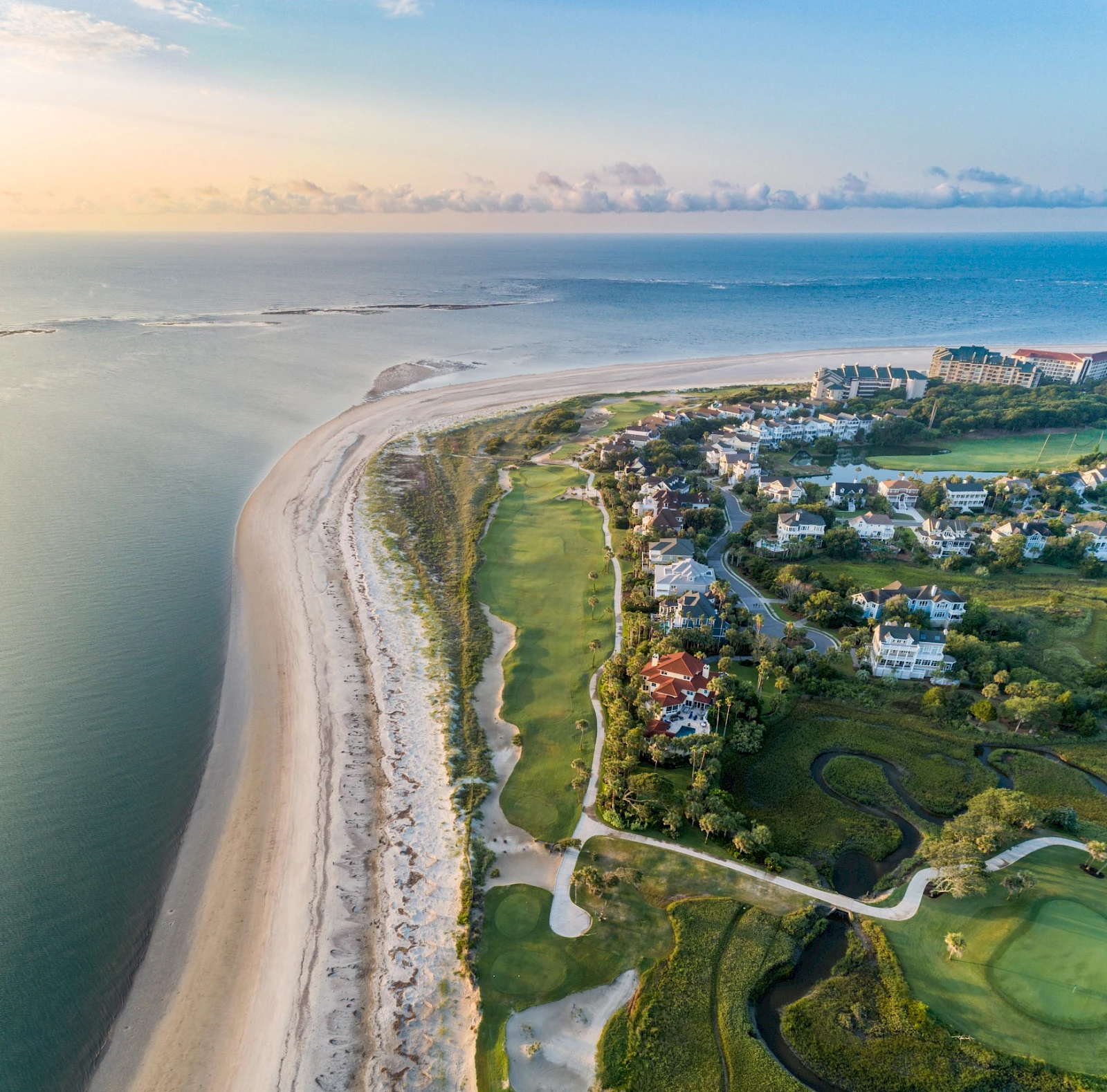 Aerial view of a coastal resort town: sandy beach, ocean on the left, residential buildings and greens on the right, golf course visible along the shore.