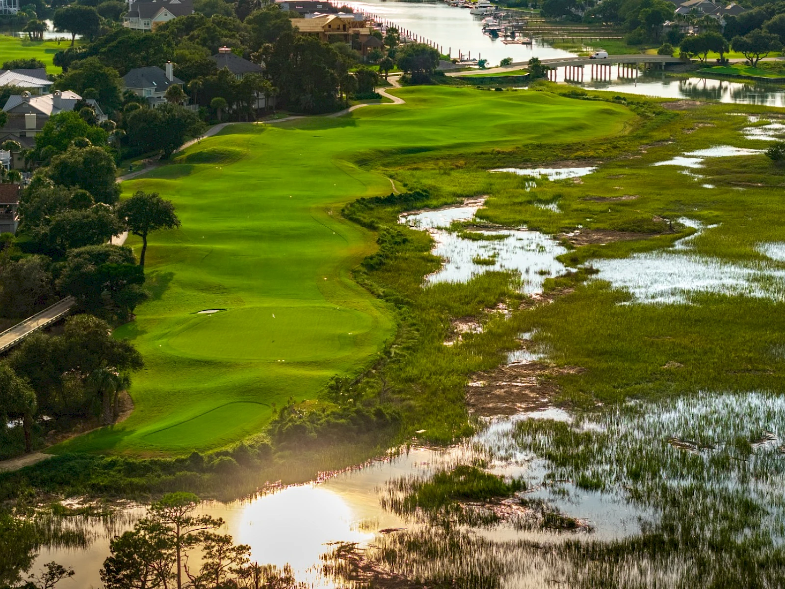 Aerial view of a lush, winding golf course bordered by houses and wetlands, with vibrant green fairways contrasting muddy, reflective water pockets.