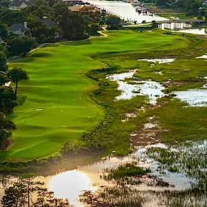 Aerial view of a lush, winding golf course bordered by houses and wetlands, with vibrant green fairways contrasting muddy, reflective water pockets.