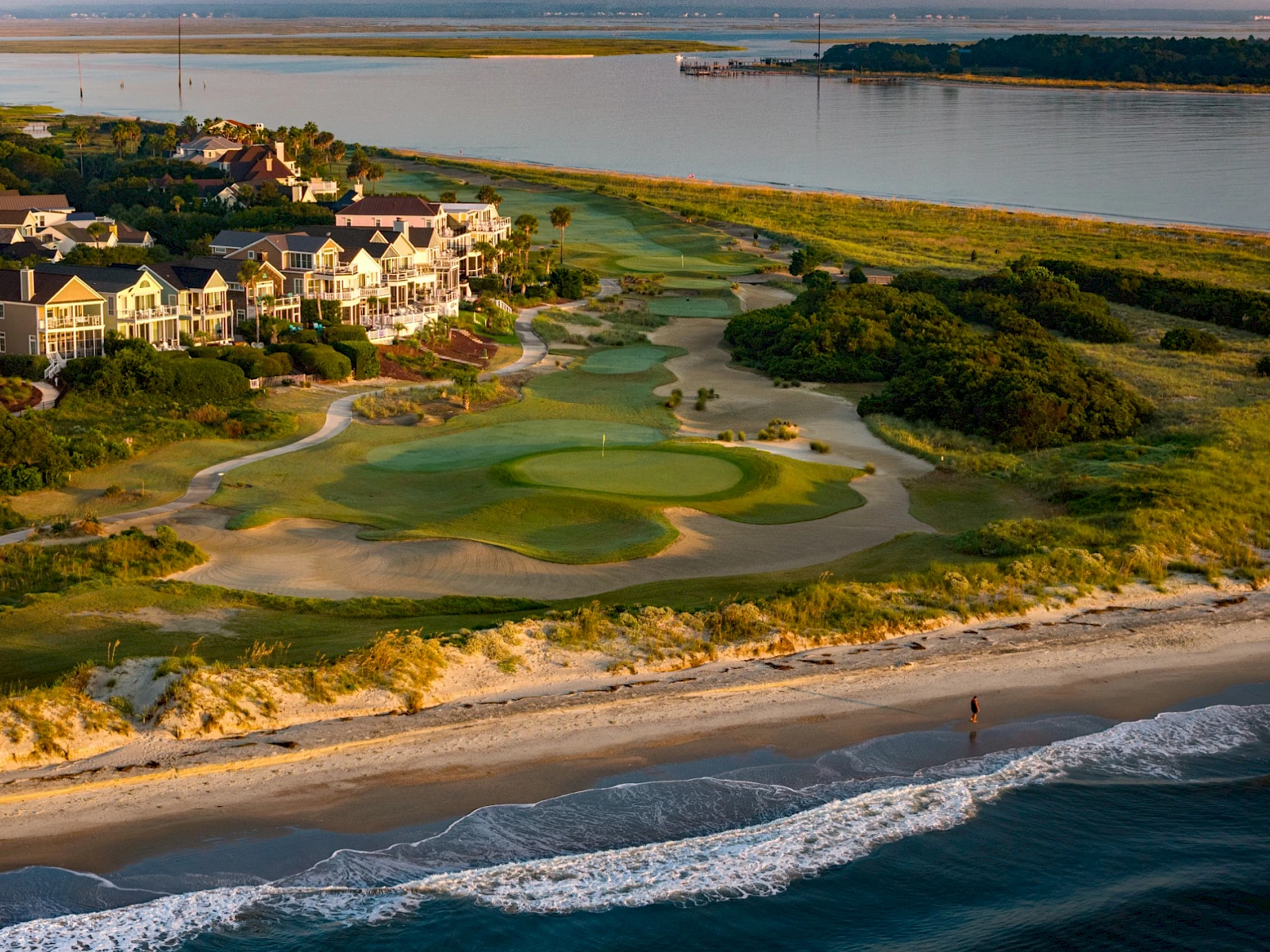 Coastal golf course with green fairways near the shore, sandy dunes, houses along the coast, and waves crashing onto the beach nearby.