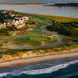 Coastal golf course with green fairways near the shore, sandy dunes, houses along the coast, and waves crashing onto the beach nearby.