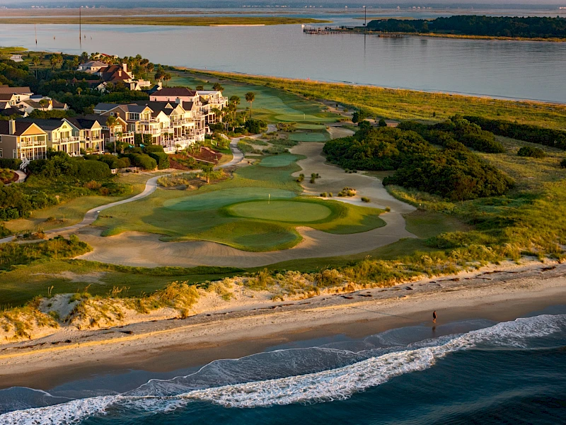 Coastal golf course with green fairways near the shore, sandy dunes, houses along the coast, and waves crashing onto the beach nearby.