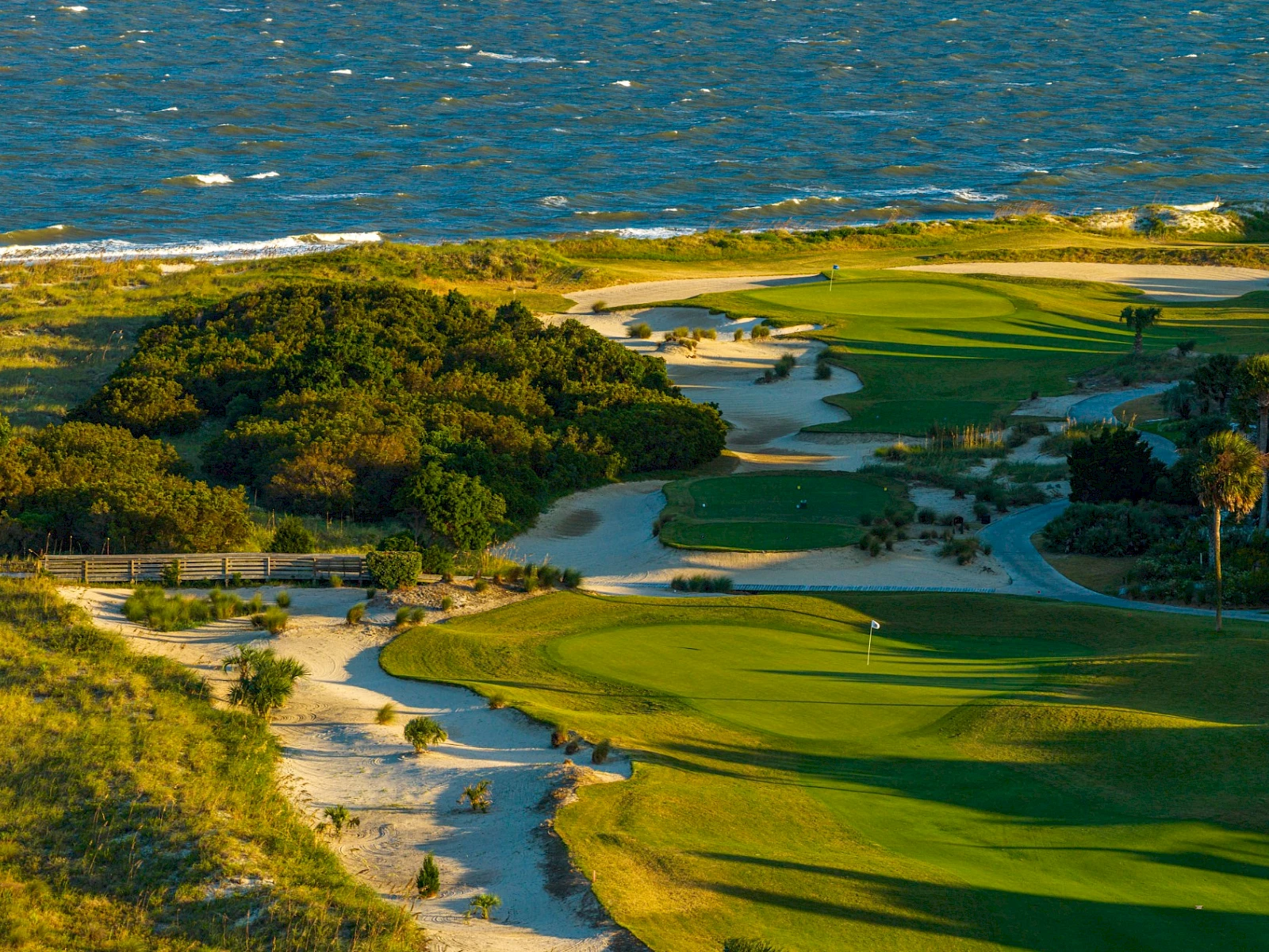 A coastal golf course with winding fairways, sand traps, and shrubs, perched above the sea on a sunny day.