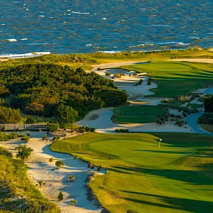 A coastal golf course with winding fairways, sand traps, and shrubs, perched above the sea on a sunny day.