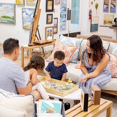 A family reads and colors together in a cozy, art-filled living room and storefront, sharing a sunny creative moment.