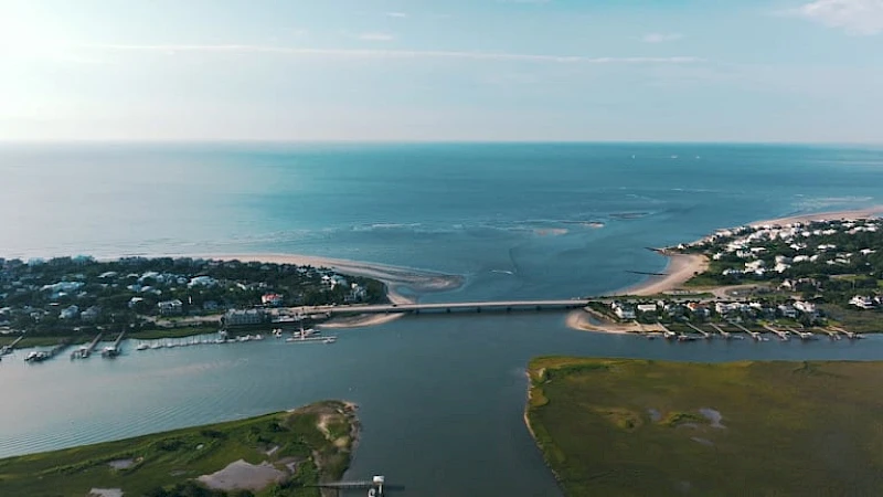 Aerial view of a coastal town with a bridge spanning a river or inlet, boats docked along shores, and expansive sea meeting the horizon.