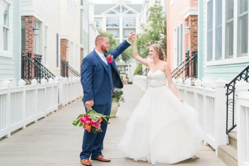 A newly married couple dances in a charming outdoor walkway, the groom in a navy suit holding a bouquet while the bride in a white gown smiles.